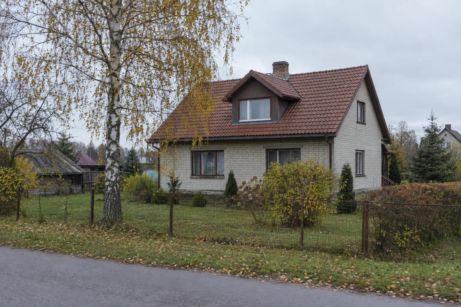 Plytinis namas su raudonų čerpių stogu ir išsikišusiu langu, priešais beržas su rudens lapais