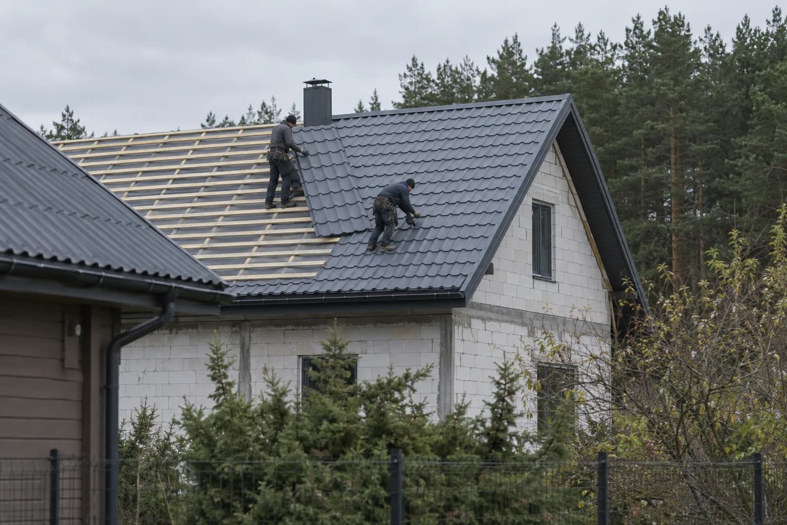 Du stogdengiai klojo tamsią metalo čerpių dangą ant pilkų blokelių namo, kitoje šlaito pusėje dar matosi grebėstai